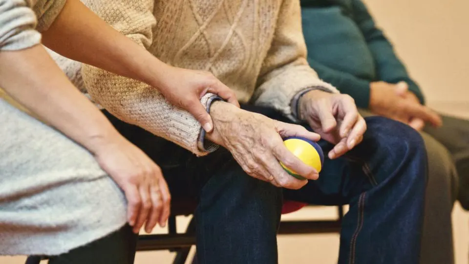 Mãos de uma pessoa idosa segurando uma bolinha de exercício, com apoio de outra pessoa